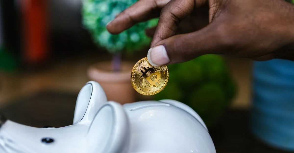 Un gros plan d'une main plaçant un bitcoin dans une tirelire blanche, symbolisant l'investissement et l'épargne.